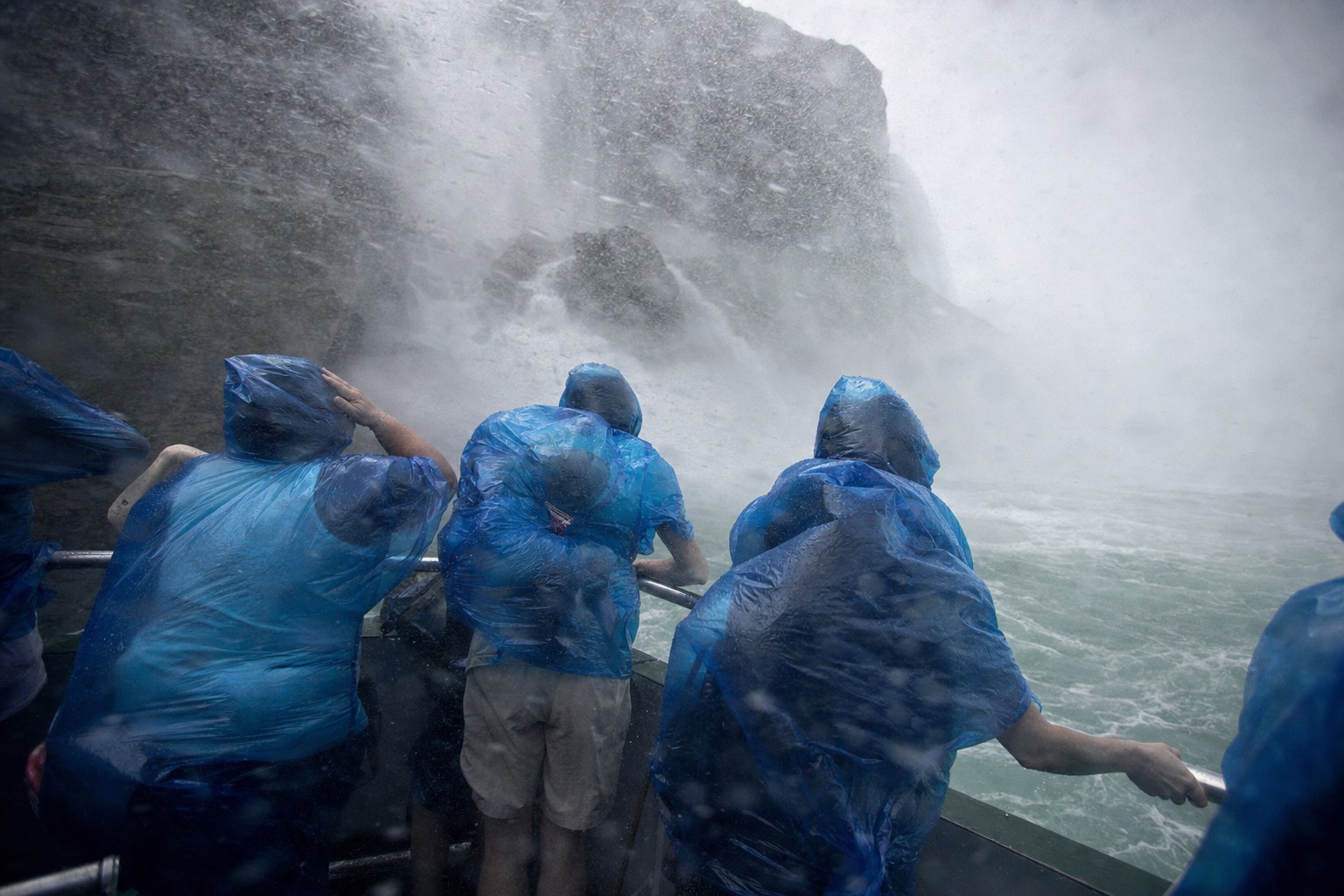Passengers at the boat rail hit by a sideways sheet of spray inside the falls basin, ponchos pressed flat by the wind