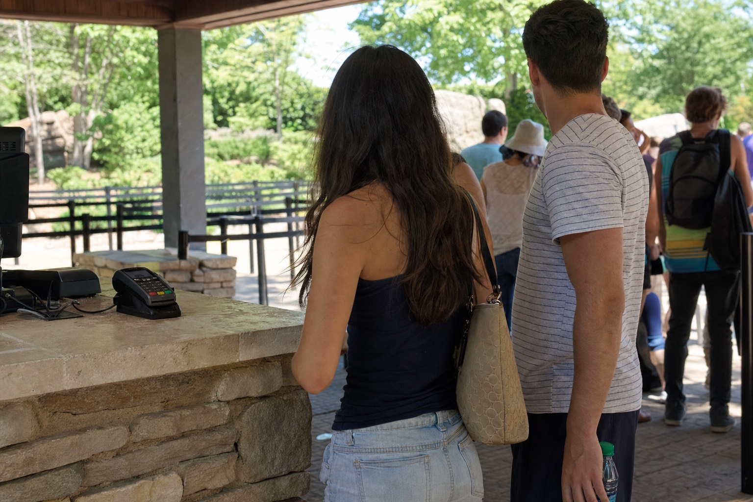 Visitors at an outdoor ticket plaza window on a bright morning, card terminal on the counter